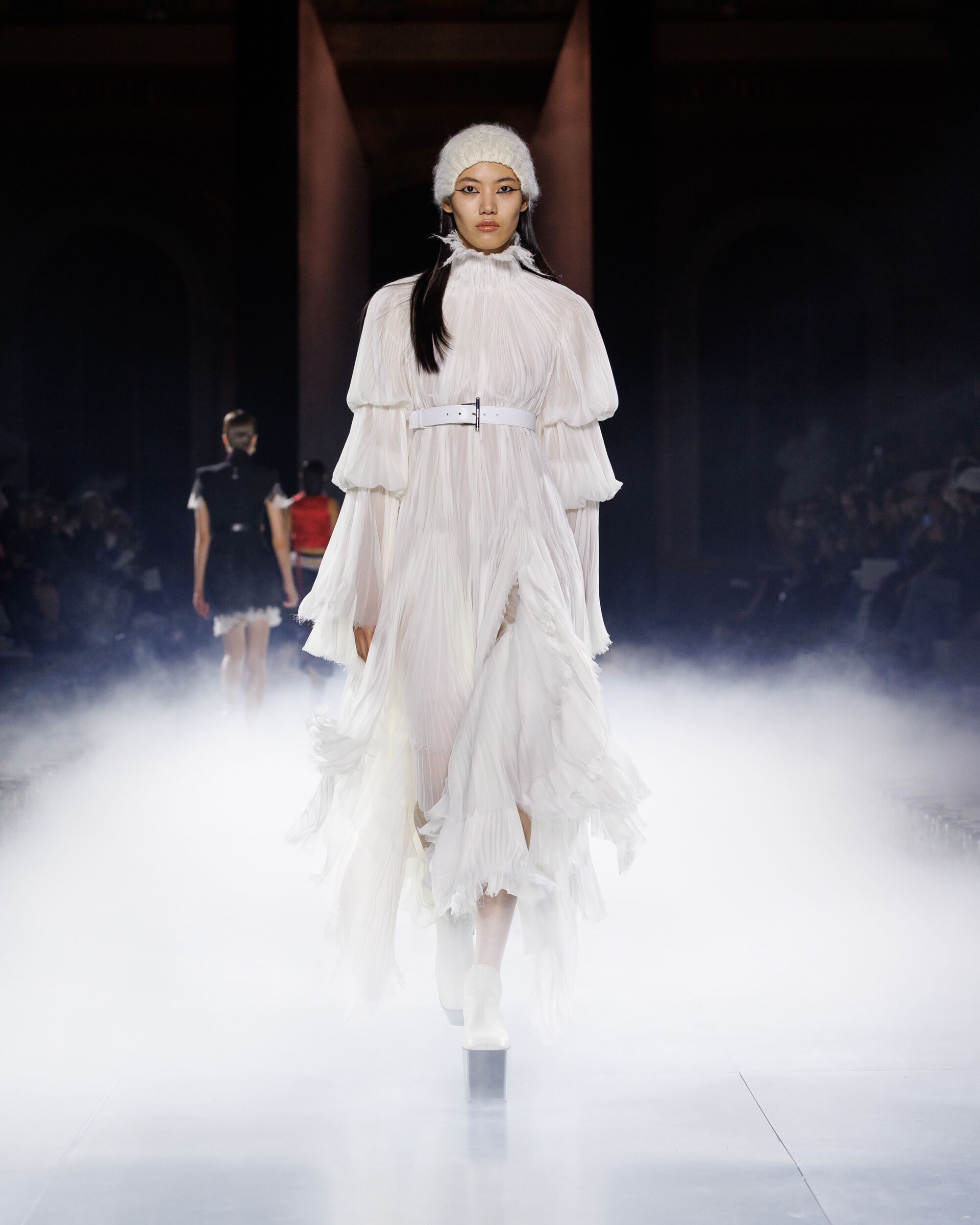 Model wearing McQueen Spring Summer 2025 Look 8 Pleated white dress with belt and knitted skull headpiece