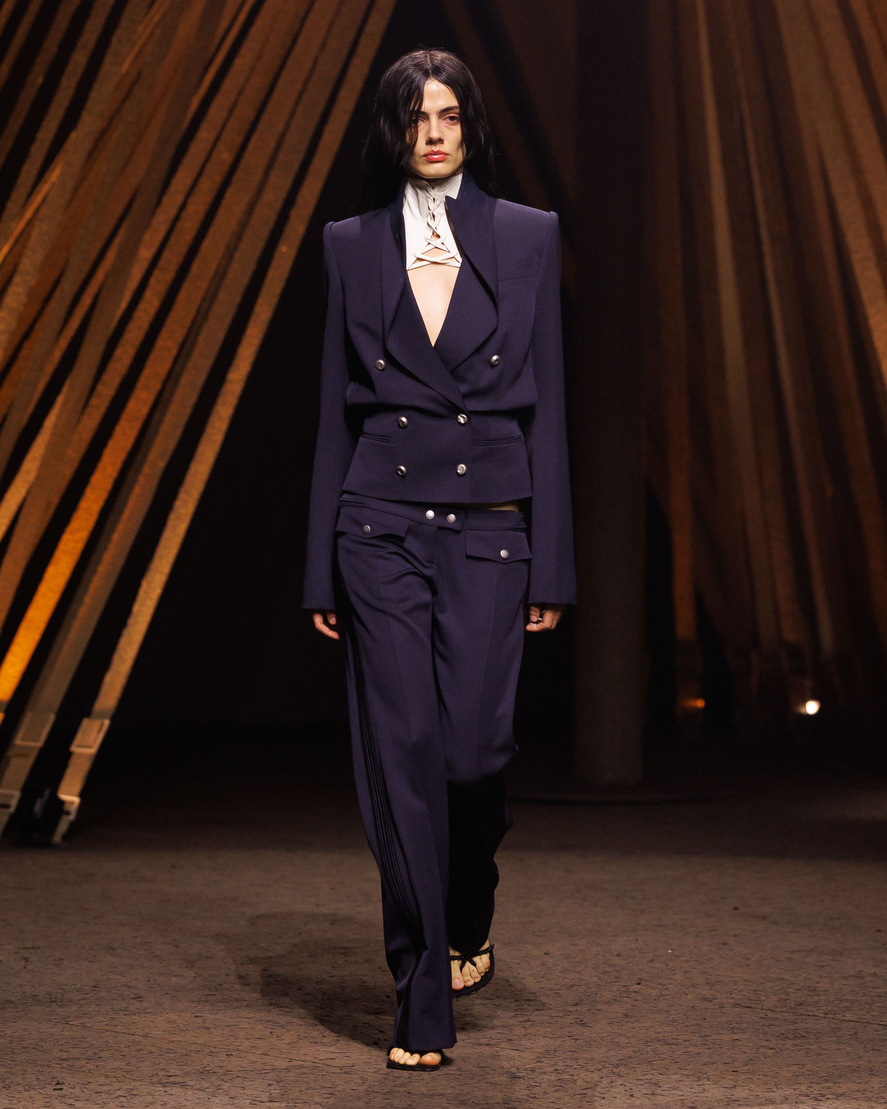 McQueen Spring Summer 2026 Look 4 double-breasted tailored jacket with pleated hourglass waist & utility bumsters in navy grain de poudre. High-neck laced harness in ivory cotton twill. Beaded sandals in black suede with horn-shaped heels.