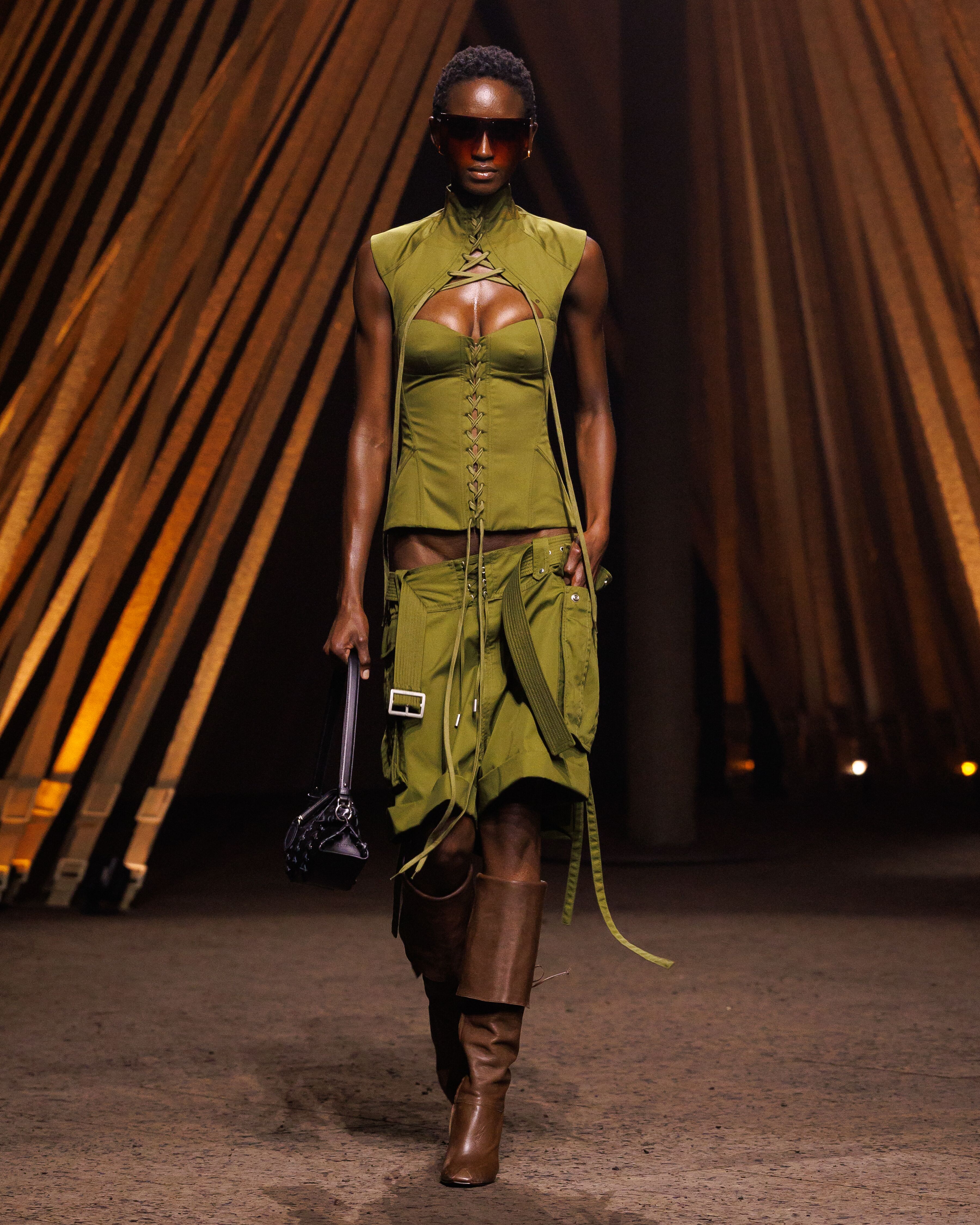 McQueen Spring Summer 2026 Look 31 Harness corset top in olive cotton twill with lacing detail. Corgo shorts in olive cotton twill with corset lacing detail. Manta clutch in black leather with corset lacing detail.
